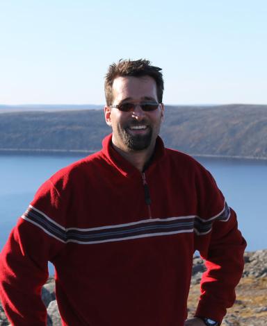 Guillaume St-Onge portant un chandail rouge avec des rayures et des lunettes de soleil, posant devant le cratère des Pingualuit au Québec qui est d’un bleu limpide