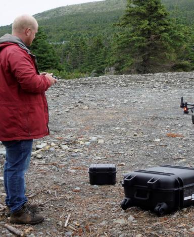 A man in a red jacket holds the remote control as a drone takes off from a rubble-strewn site. 