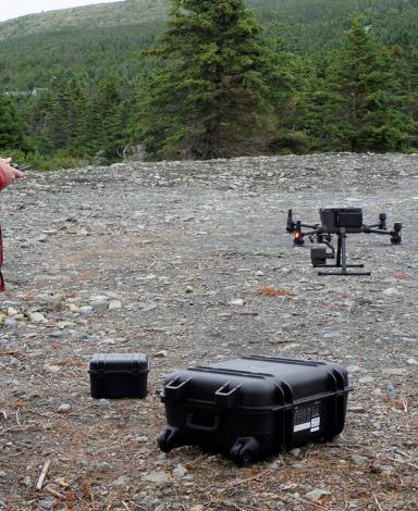 Un homme vêtu d’un manteau rouge tient une télécommande alors qu’un drone décolle d’un site jonché de débris.