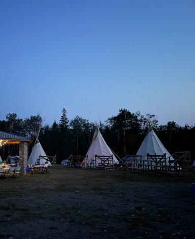 An outdoor scene at dusk with a stone pavilion and several tipis arranged in a circle with bleachers.