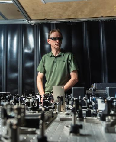 David Jones porte des lunettes de protection laser, debout derrière un prototype posé sur son plan de travail de laboratoire.