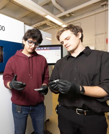 Two students wearing disposable gloves each hold a small metal object as they stand in front of a large TRUMPF TruPrint 2000 machine.