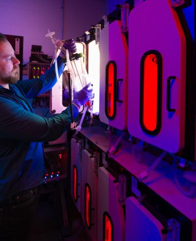 A scientist inside a lab pulls a large, clear cylinder from inside one of several illuminated chambers.