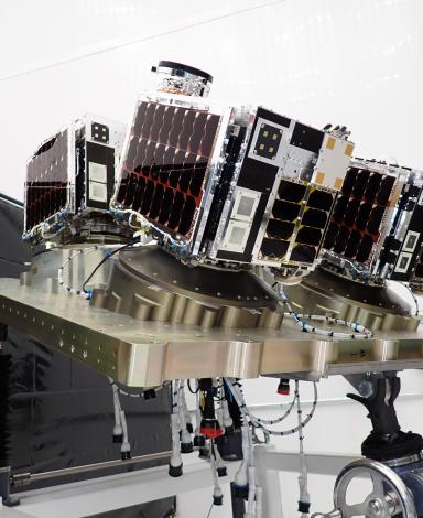 Vue dans un laboratoire de trois satellites, chacun de la taille d’une boîte à chaussures, montés sur une plateforme métallique.