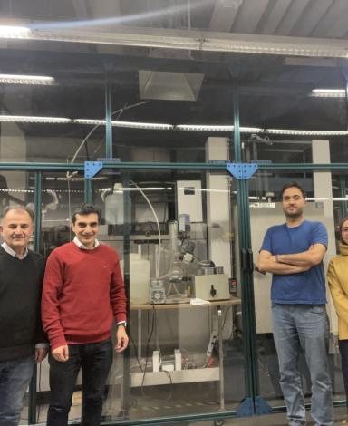 Four researchers stand in front of a large, glassed-off section of a lab filled with experimental equipment.