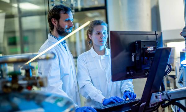 Two people wearing lab coats and gloves look at a computer screen