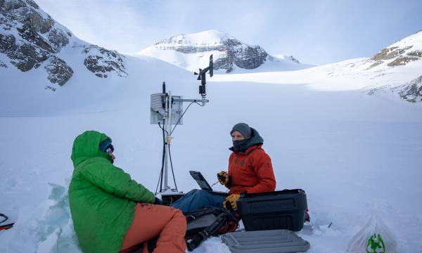 Deux personnes en habits d’hiver sont assises dans la neige en montagne entourées de matériel de recherche.