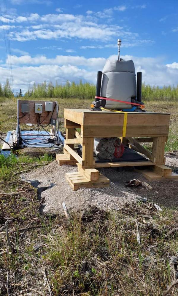 A machine the size of a mini-fridge sits on an elevated wooden palette in a field of low grass with a forest of small evergreens in the background.