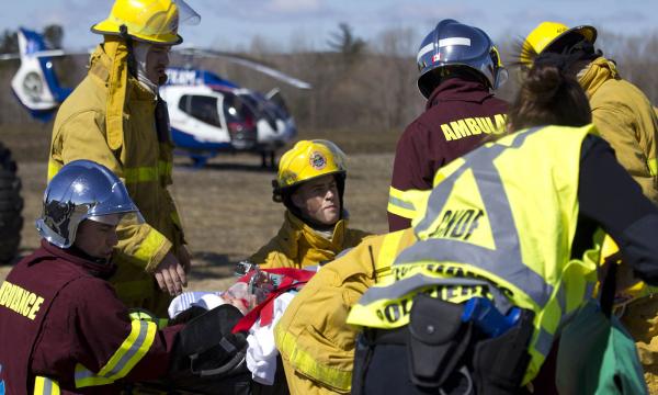 Simulated disaster scene showing a person lying on a stretcher and six other people providing care. The responders are wearing vests with the word “ambulance” on them or fluorescent yellow sleeveless vests, as well as helmets. 