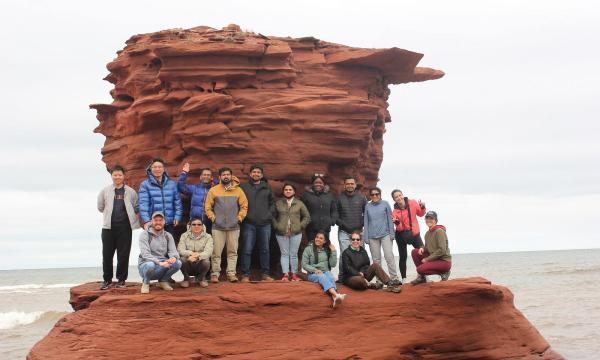 A group of researchers pose in front of a huge red rock formation shaped like a teacup by the ocean in Prince Edward Island.