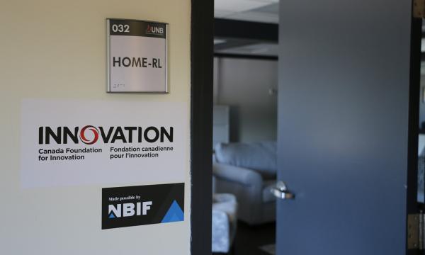 Une porte ouverte débouche sur une pièce où trônent des canapés. À côté de la porte se trouvent trois affiches; l’une porte le nom du laboratoire (HOME-RL), et les deux autres, le logo de la Fondation canadienne pour l’innovation et celui de la Fondation de l’innovation du Nouveau-Brunswick.