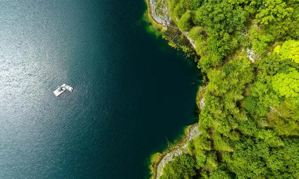 An aerial view of a boat by a raft in a shimmering lake near a green, wooded shoreline.