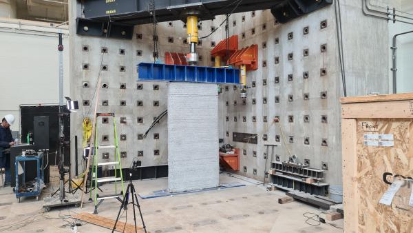 A steel beam is pressed onto the top of a 3D-printed wall in a large lab space.