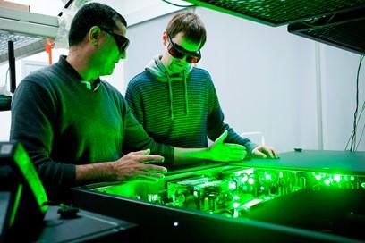 Two persons wearing glasses are bathed in a fluorescent green light emanating from a measuring instrument using lasers.