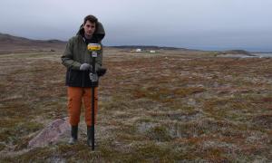 A researcher holding a pole with GPS equipment stands on a rough, grassy field with ocean in the background.