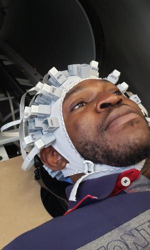 A person wearing a cap covered in sensors lies on their back preparing to enter the cylinder of a medical imaging machine.