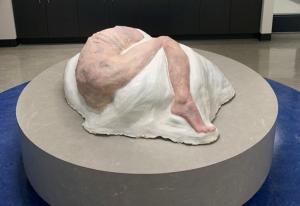 Une sculpture représentant un corps humain couché sur le côté et roulé en boule repose sur un socle circulaire.