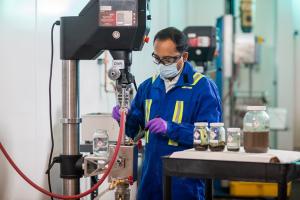 A person in blue overalls, with a mask, glasses and purple gloves is operating the Batch Extraction Unit (BEU). On a work bench next to them are four jars of different sizes filled with soil samples.