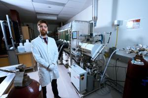 Debout dans son laboratoire, Arthur Situm porte des lunettes de protection, une blouse blanche et des gants. À ses côtés se trouvent une boîte à gants, une bouteille de gaz et divers équipements.