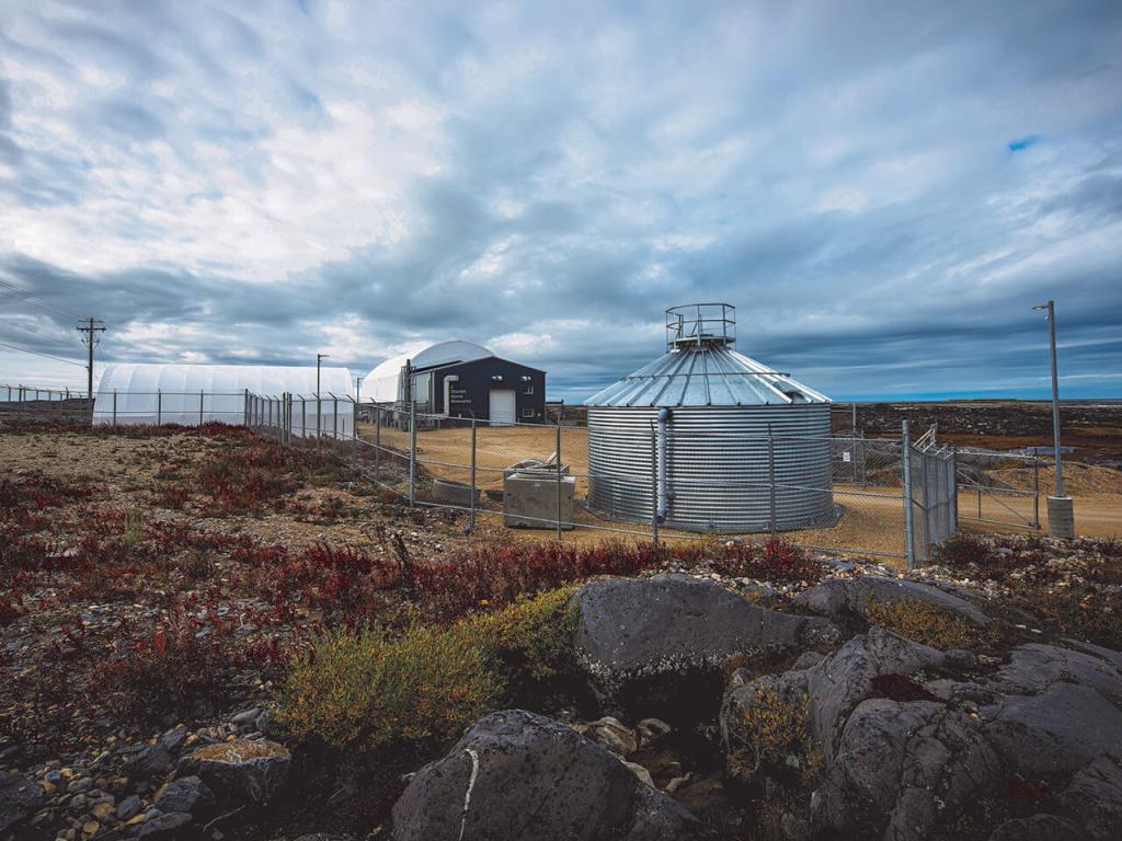 A rocky outcrop with low brush in the foreground with a silver cylindrical building in the middle ground behind a fence and a large domed structure in the background