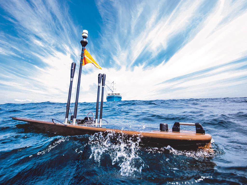 A vessel the size and shape of a surfboard with an apparatus attached glides on the surface of the ocean with a ship in the background.