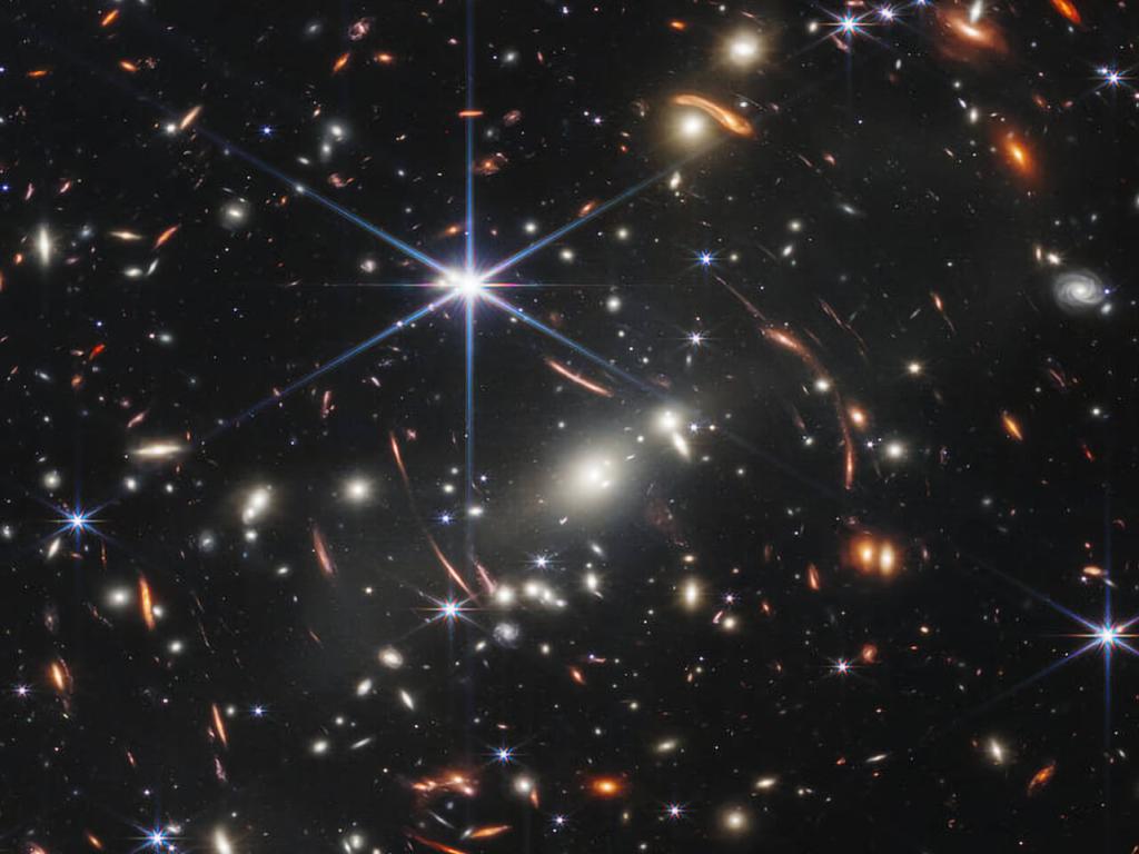 Distant galaxies appear as bright glowing spots in this Webb telescope image, with some smeared by gravitational lensing; foreground stars appear bright with six-pointed diffraction spikes, owing to the shape of Webb's mirrors. 