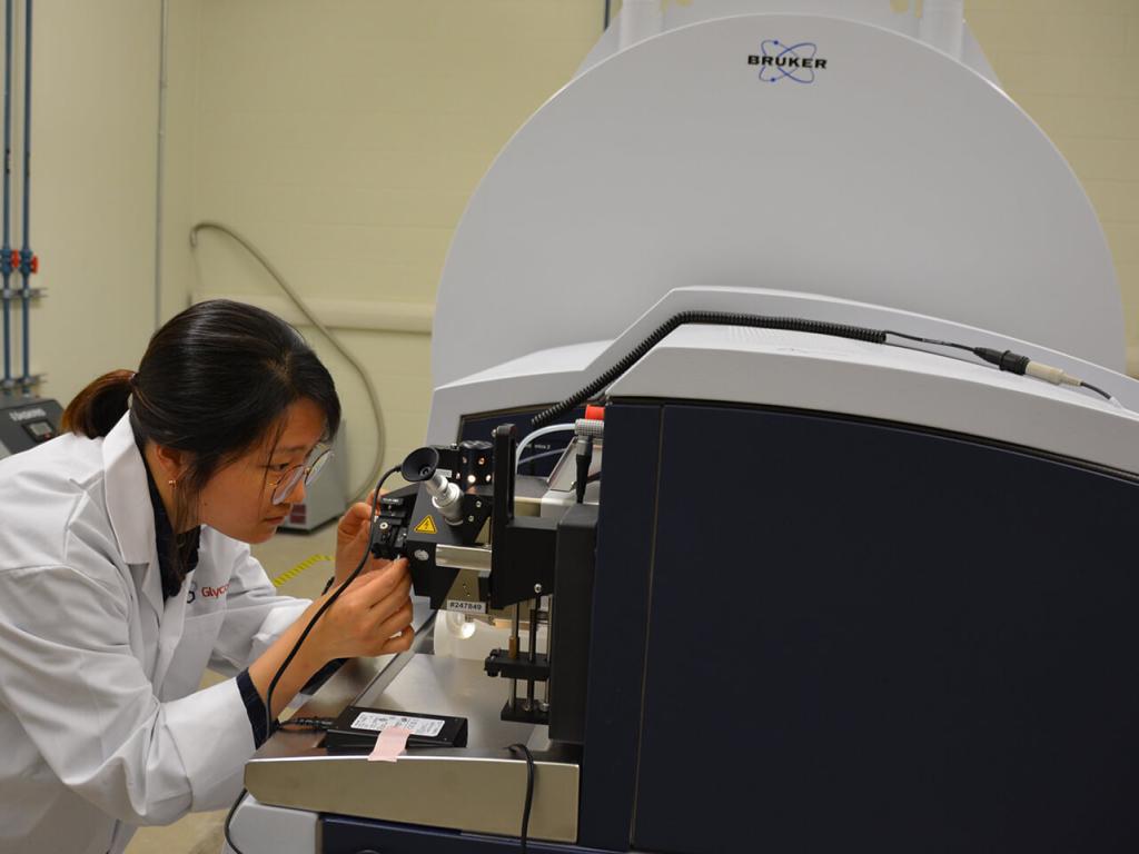 A person in a lab coat looks closely at an apparatus that is part of a black and grey machine in a lab.