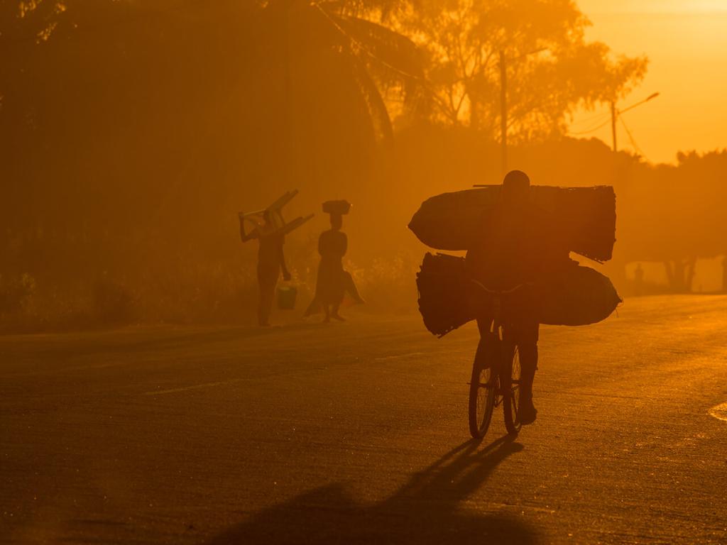 Photo dans un dégradé de couleur jaune uniquement d'une personne sur un vélo portant une lourde charge à contre-jour dans un lever de soleil rendant l’image brumeuse.