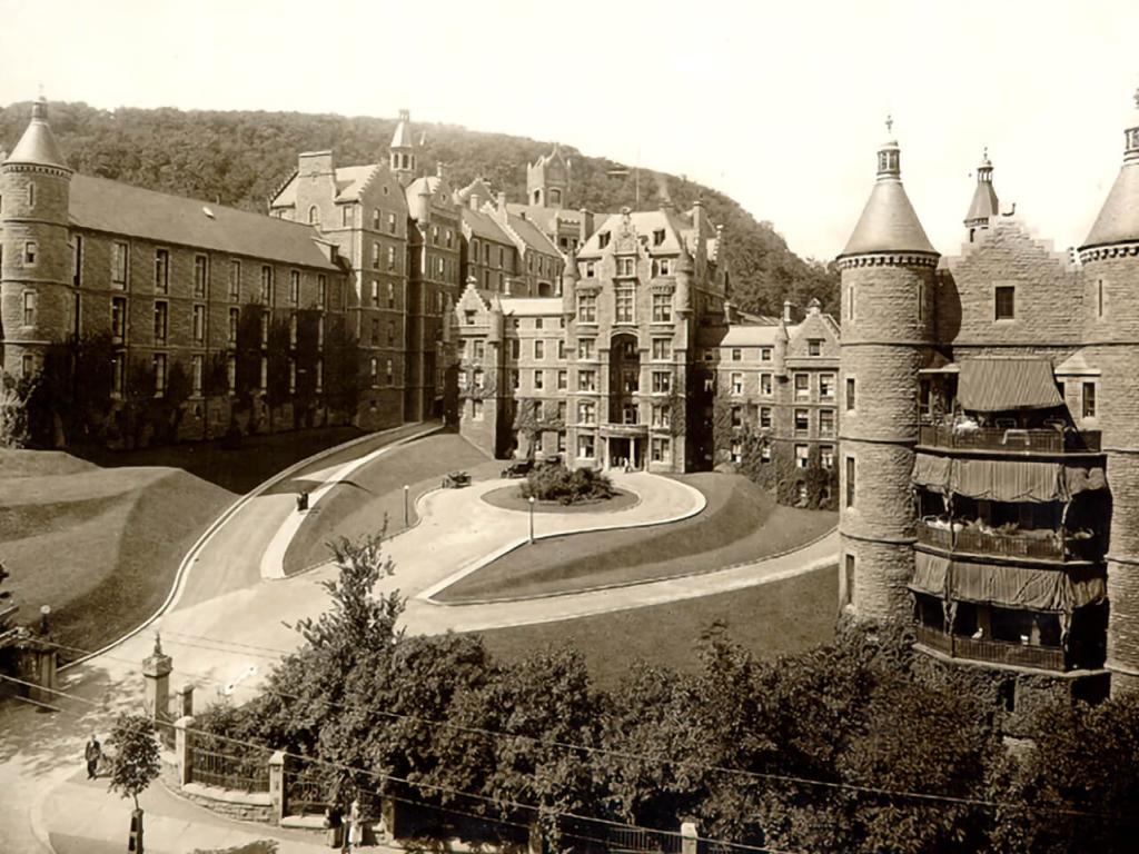 A grey and white photo of a multi-storeyed building with turrets at each corner and a large circular driveway in front.