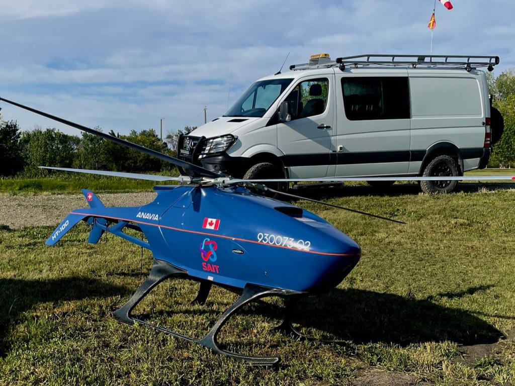 A drone resembling a small blue helicopter sits on a grassy area in the foreground with a white van parked on a driveway in the background.