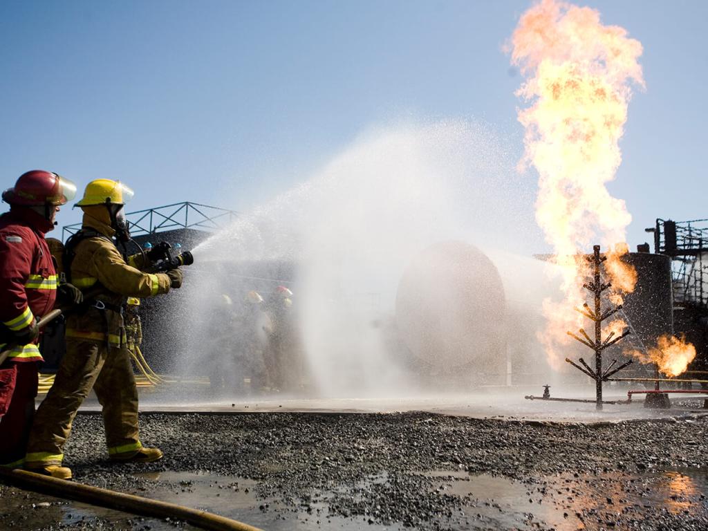 Photo of a fire simulation exercise. Two people wearing fire coats aim a fire hose at a burning building. 