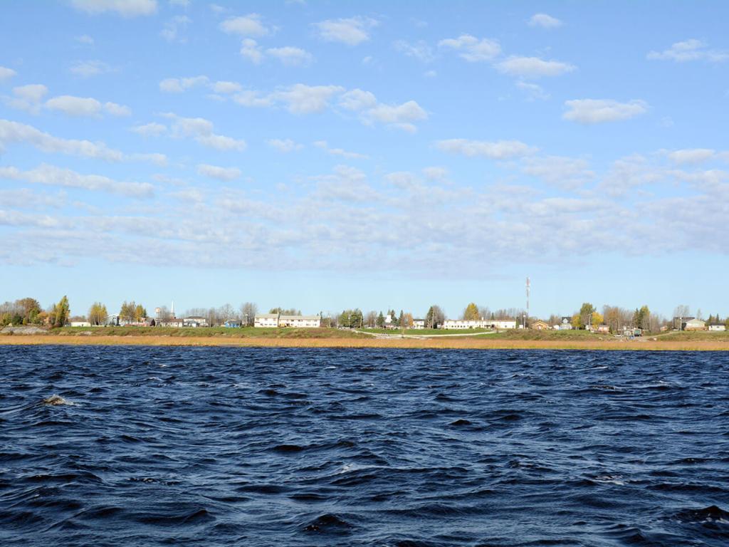 Low buildings and homes along a flat shoreline with water in the foreground. 
