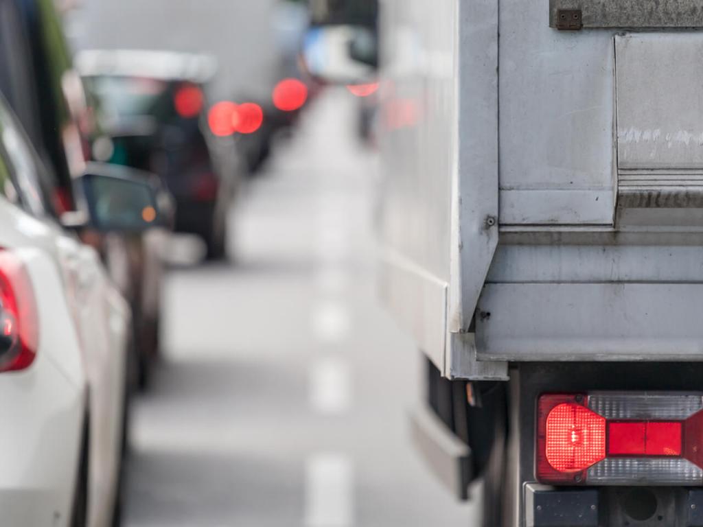 A close-up photo of taillights of a car and a transport truck at the back of a long line of traffic.