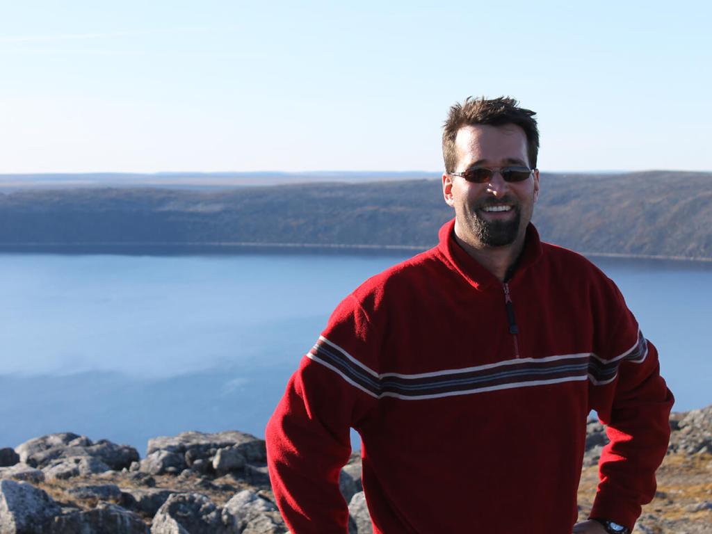 Guillaume St-Onge is wearing a striped red sweater and sunglasses, posing in front of the Pingualuit crater in Quebec, full of clear blue water.