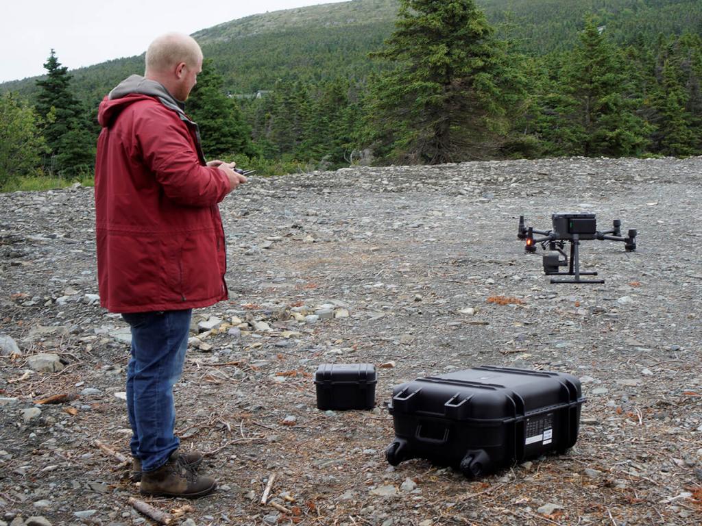 A man in a red jacket holds the remote control as a drone takes off from a rubble-strewn site. 