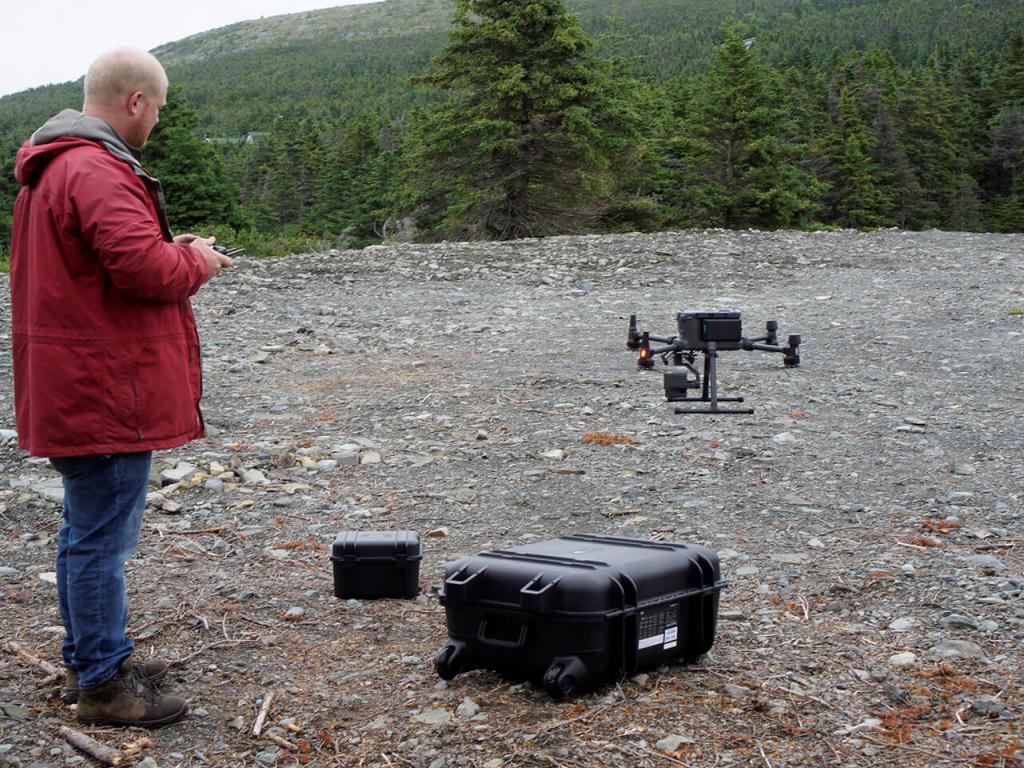 Un homme vêtu d’un manteau rouge tient une télécommande alors qu’un drone décolle d’un site jonché de débris.