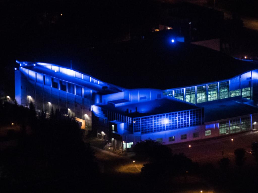 Vue nocturne, en plongée, d’un grand bâtiment dont les murs sont éclairés d’une vive lumière bleue.