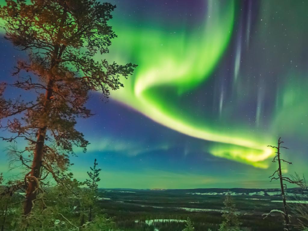 Un tourbillon de lumière verte dans le ciel au-dessus d’un paysage peu boisé.