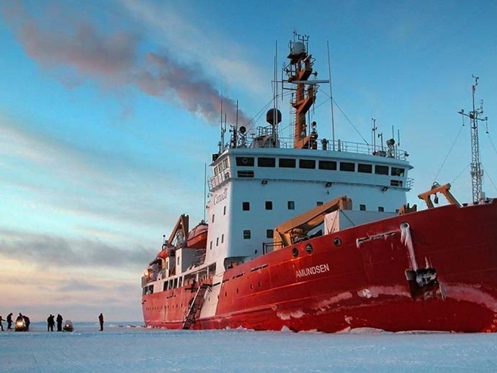 A small group of people stand on a vast expanse of ice, dwarfed by the red and white ice breaker beside them. 
