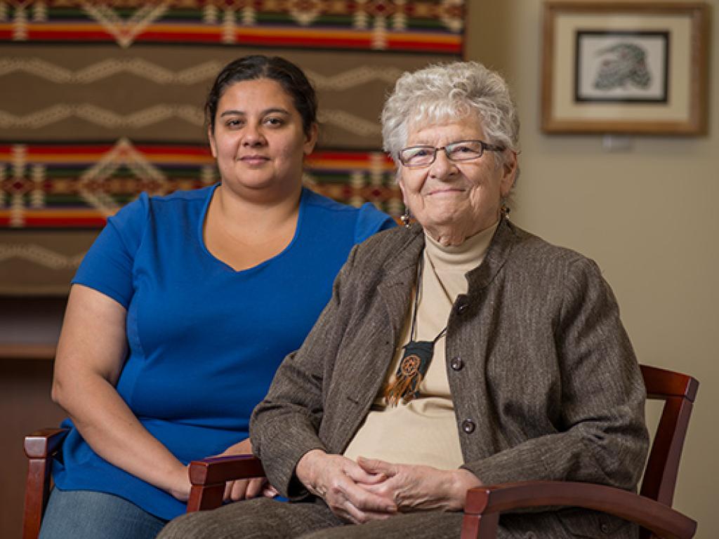 A young woman in blue and an older woman wearing a brown jacket and necklace that looks like a pouch with leather fringe sit side-by-side in a room in front of a patterned wall hanging