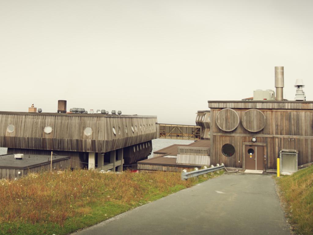 Un bâtiment en bois semblable à un bateau, muni de hublots ronds se dresse sur un littoral; le rivage opposé est couvert de brouillard.