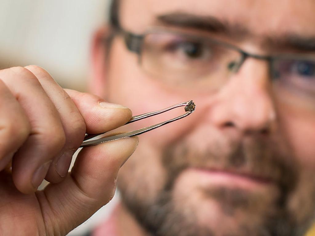 A man uses tweezers to hold up a small diamond. The diamond is in focus, blurring out the man’s face.