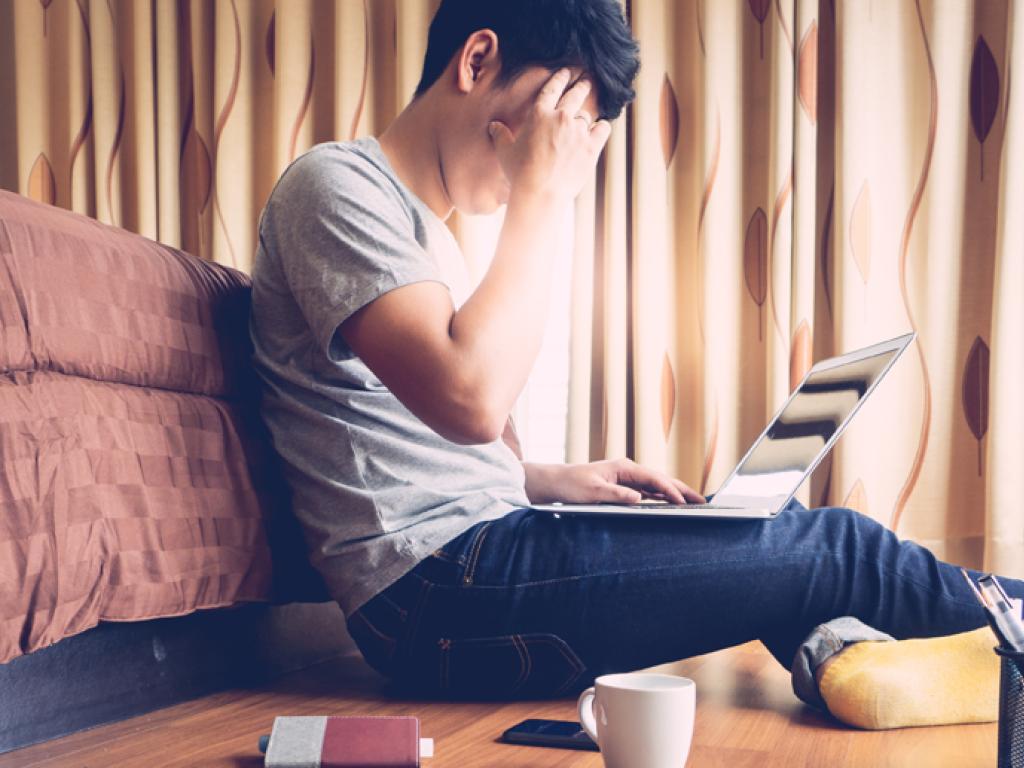 A person puts a hand to their forehead while sitting on the floor and looking at a laptop 