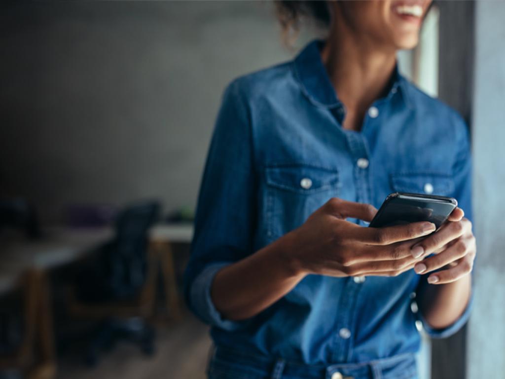 A woman, who is blurred out in the background, holds a mobile phone in the foreground