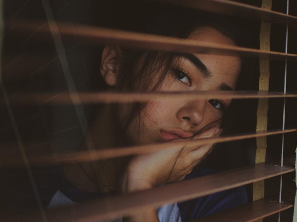 Young woman resting her face in her hand stares out a window through open shutters looking bored