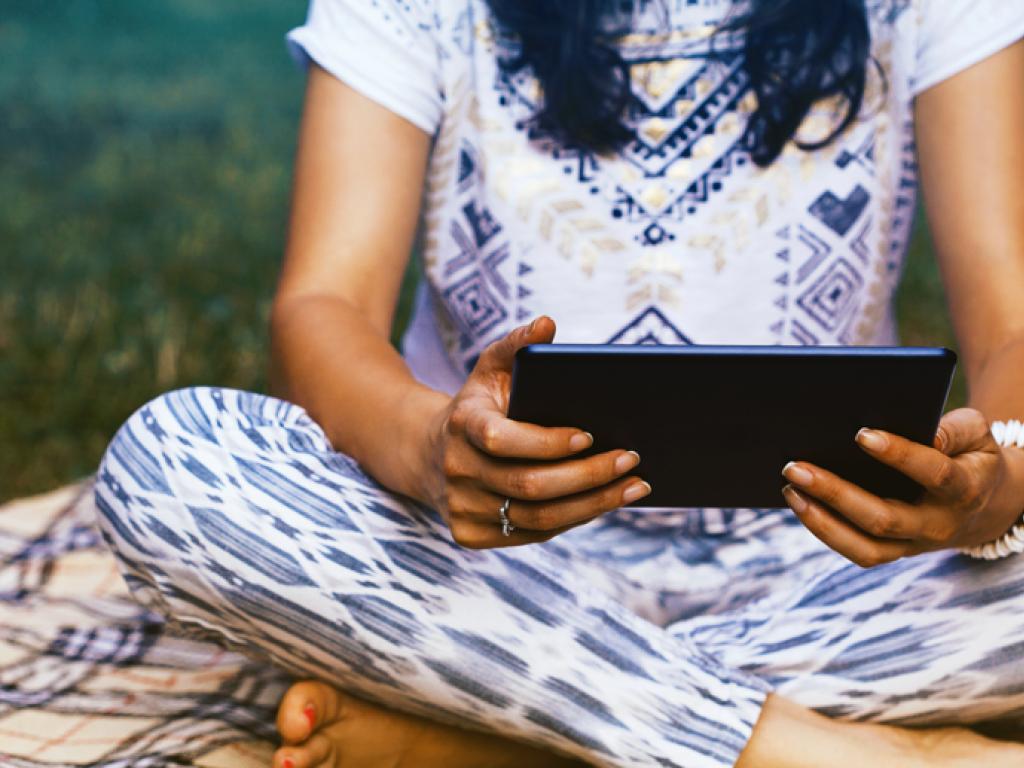 A woman sits cross-legged in the grass, holding an electronic tablet.
