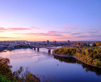Photo de la rivière des Outaouais vue de la colline du parlement au coucher du soleil. On y voit le musée canadien de l’histoire, la ville qui s’étend, le pont Alexandra qui enjambe la rivière ainsi que de la verdure au premier plan.