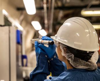 A person wearing a hard hat and gloves looks through a device while standing in a lab