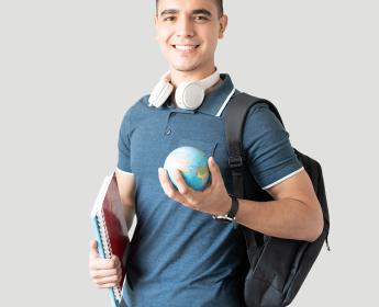 A young person standing in front of a grey background holding a miniature globe and 2 notebooks.