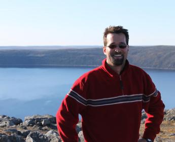 Guillaume St-Onge portant un chandail rouge avec des rayures et des lunettes de soleil, posant devant le cratère des Pingualuit au Québec qui est d’un bleu limpide